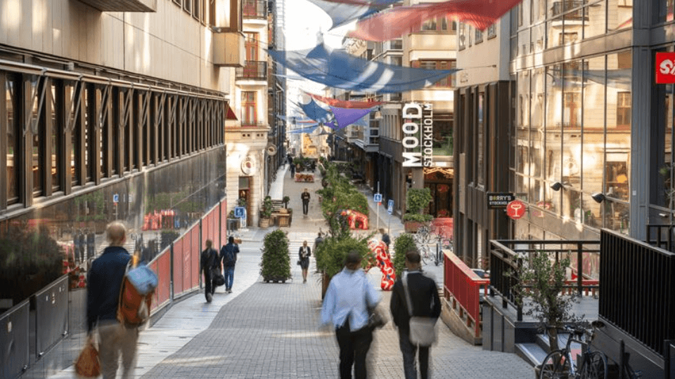 Fotgängare på en sluttande gågata i Stockholm med "MOOD Stockholm"-skylten och färgglada segel uppspända mellan fasaderna.