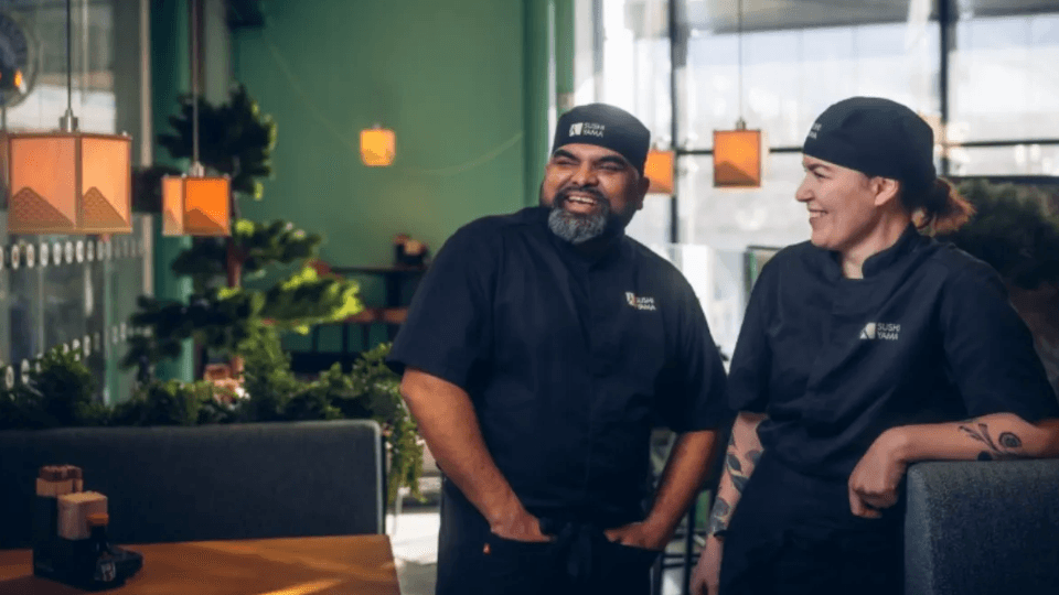 Två glada kockar i svarta arbetskläder och mössor från Sushi Yama står och samtalar i en restaurangmiljö.