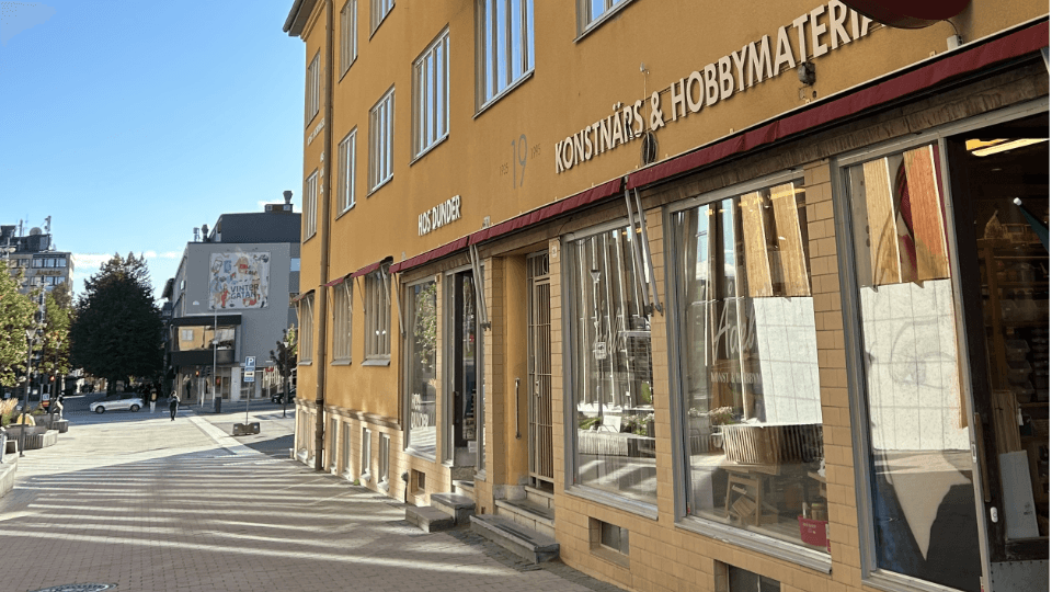 En vy över en stenbelagd gata och en gul husfasad. I bottenvåningen syns stora skyltfönster. I bakgrunden syns en blå himmel och människor som rör sig på gatan.