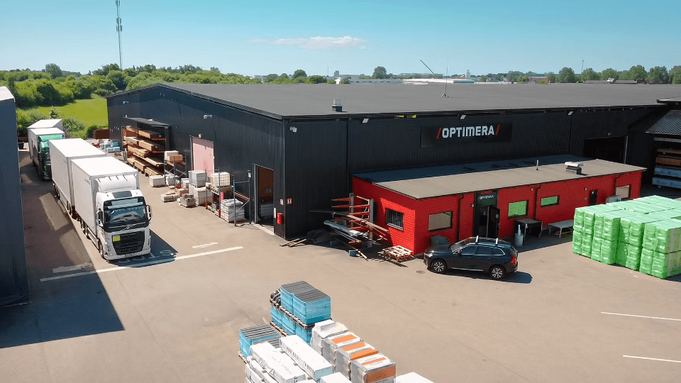 Flygfoto över en Optimera byggvaruhandel med en stor svart lagerbyggnad, en röd kontorsentré, lastbilar och byggmaterial på gården.