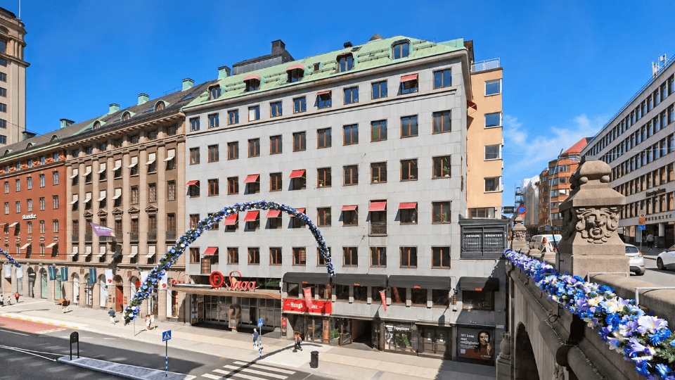 Exteriörbild i fågelperspektiv över ett stadslandskap med flervåningsbyggnader, biografen Saga, en vägbro och en blomsterbåge under klarblå sky.